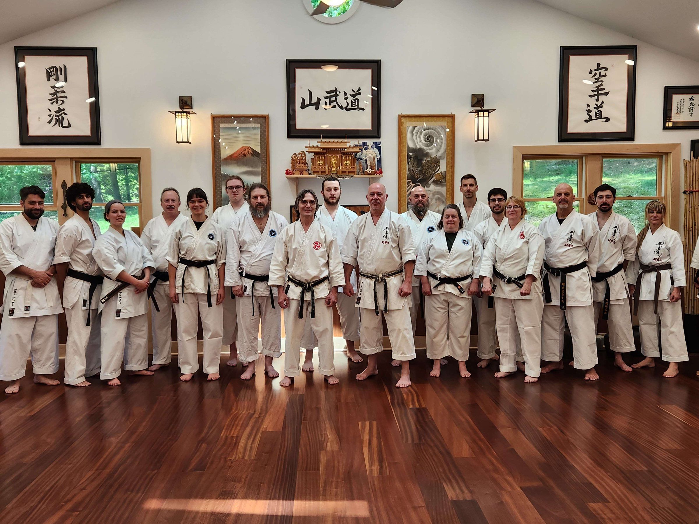 A long row of black belts in white gi at the Holmdel dojo, under framed calligraphy including 山武道.