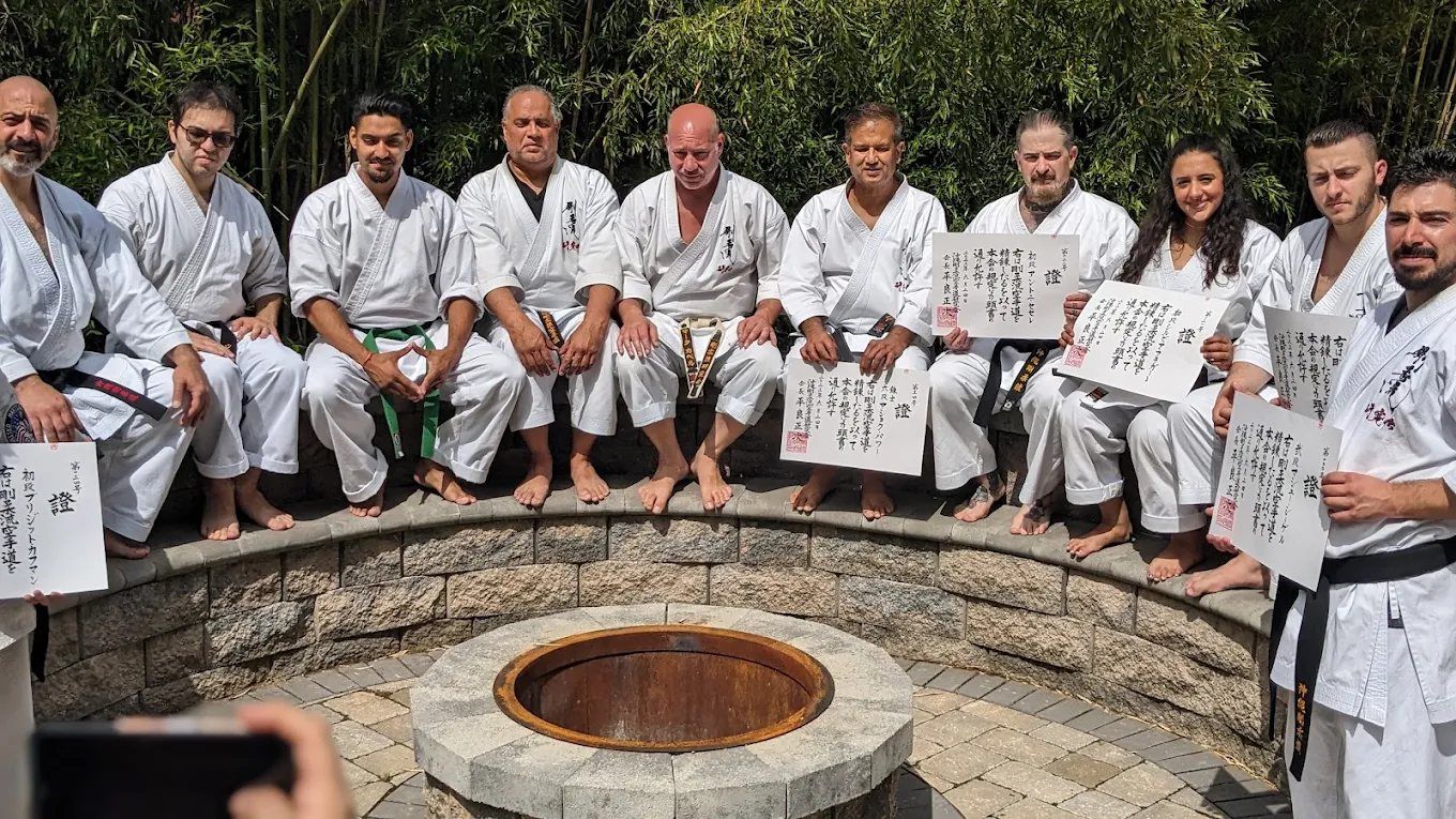 Black belts in white gi sit around a stone fire pit holding rank certificates.