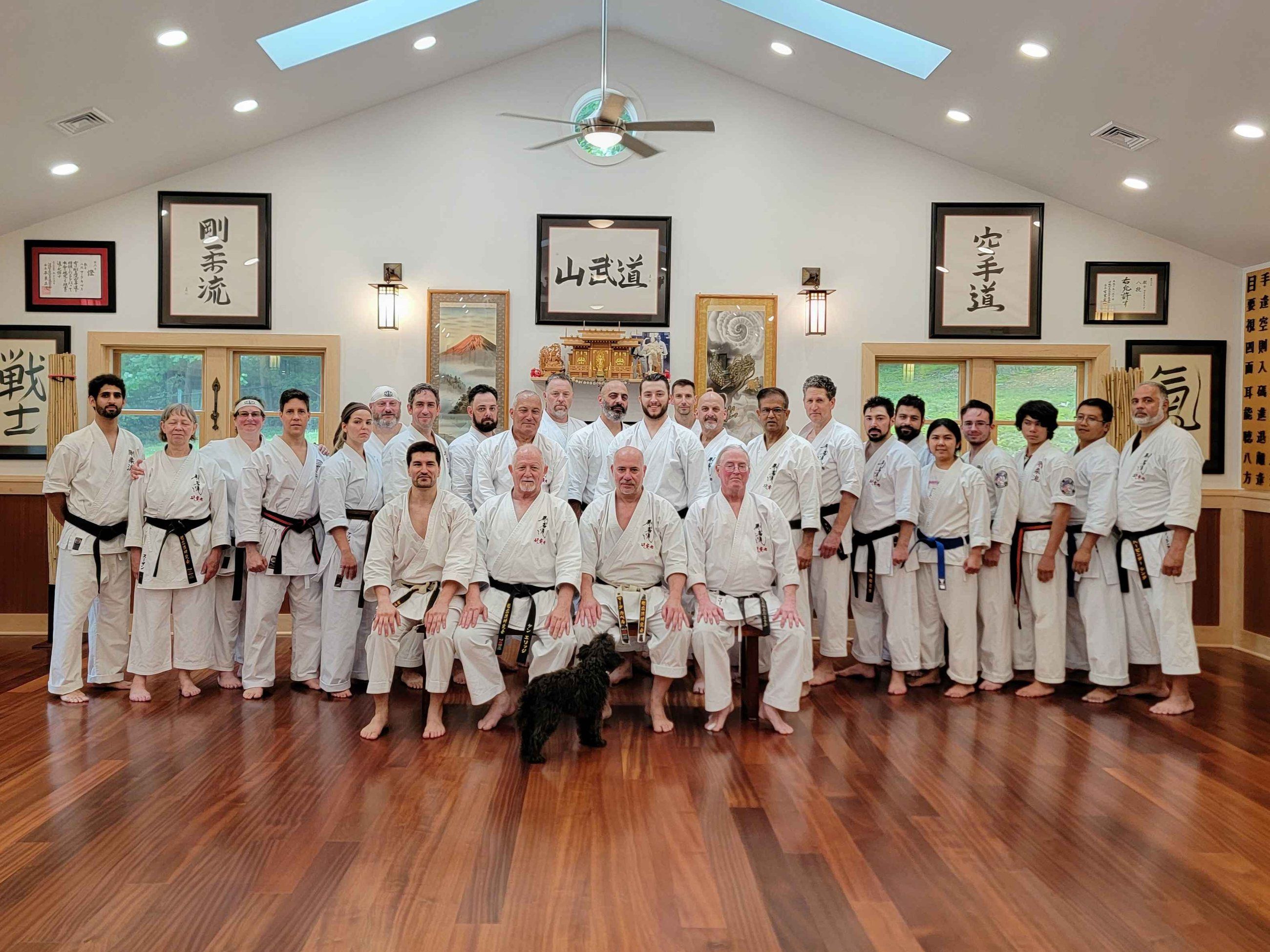 About forty karateka in white gi stand and sit in rows on the wood floor of the Holmdel dojo after a seminar, under framed calligraphy including 山武道, with a small black dog in front.