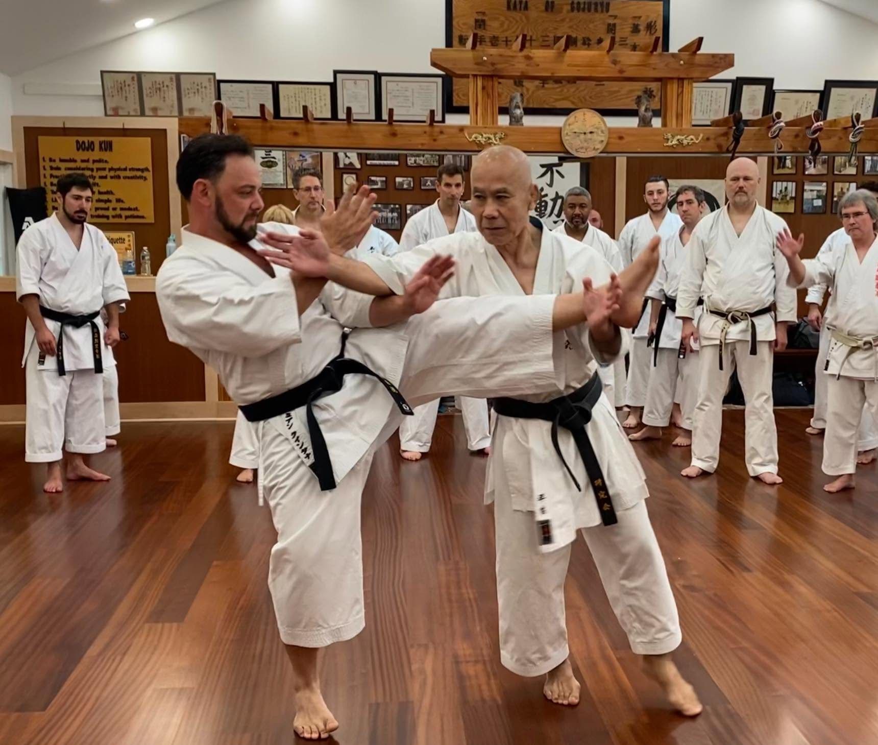 At a seminar in the Holmdel dojo, Masaji Taira Sensei blocks a kick from a black belt while a circle of students watches.