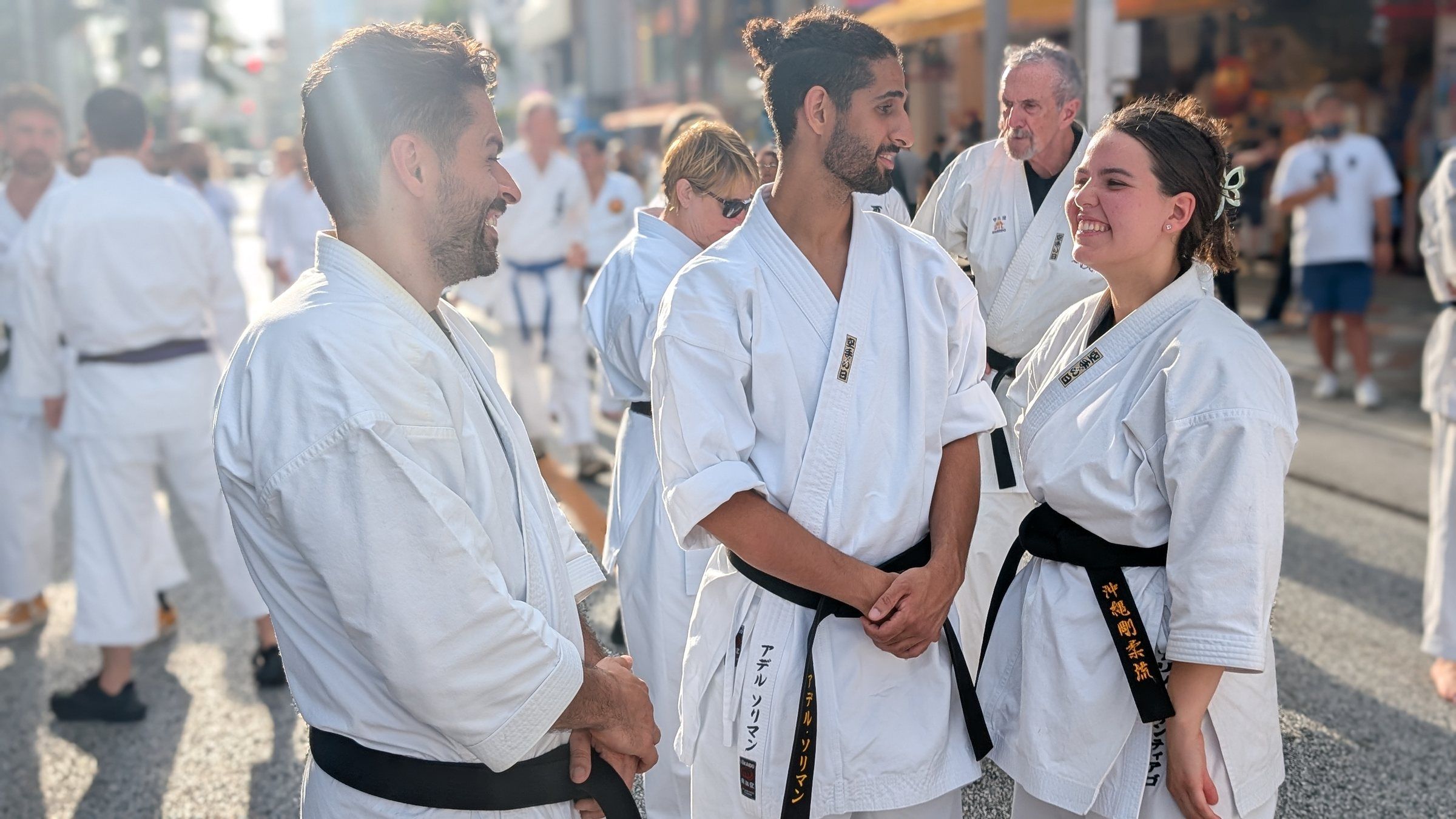 Three black belts in white gi laugh together on a sunny street in Naha during Karate Day 2025.