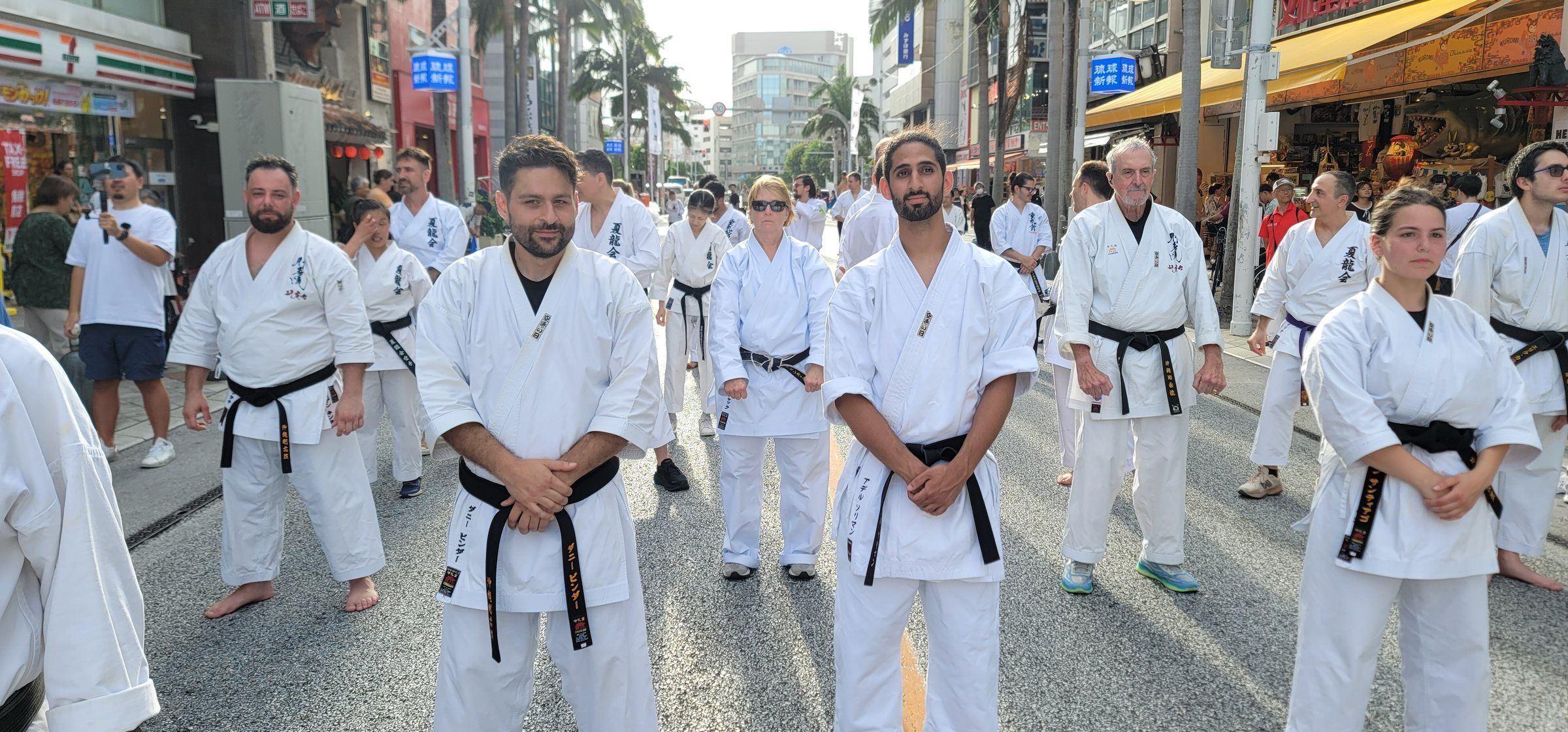 Black belts in white gi stand in rows on Kokusai-dori in Naha, Okinawa, lined with palm trees and shops.