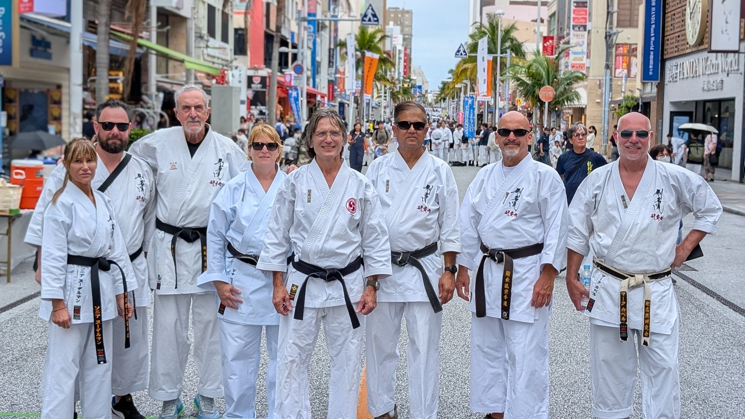 Eight black belts from the dojo in white gi stand together on Kokusai-dori in Naha, Okinawa, on Karate Day 2025.