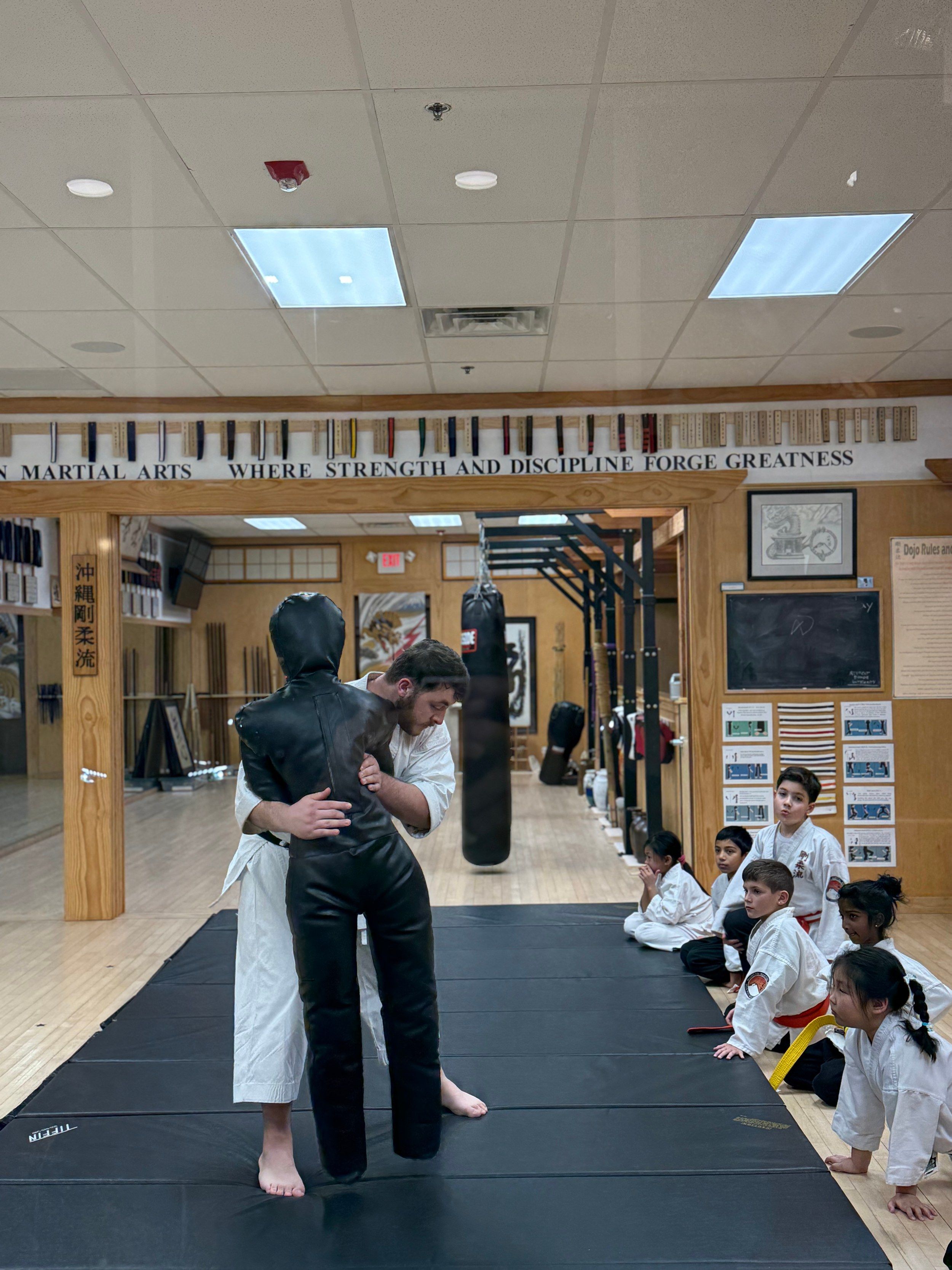 At the Old Bridge dojo, an instructor demonstrates on a grappling dummy while kids in white uniforms watch from the edge of the mat.