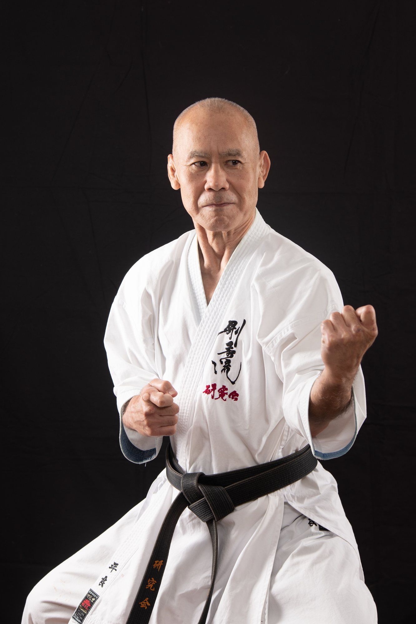 Studio portrait of Masaji Taira Sensei in a white gi and black belt, in a fighting stance against a black background.